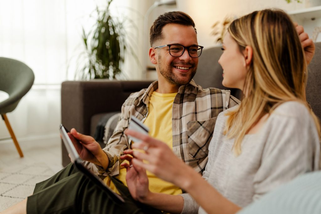 Photo of a cheerful young loving couple at home on floor near sofa using laptop computer and holding credit card.