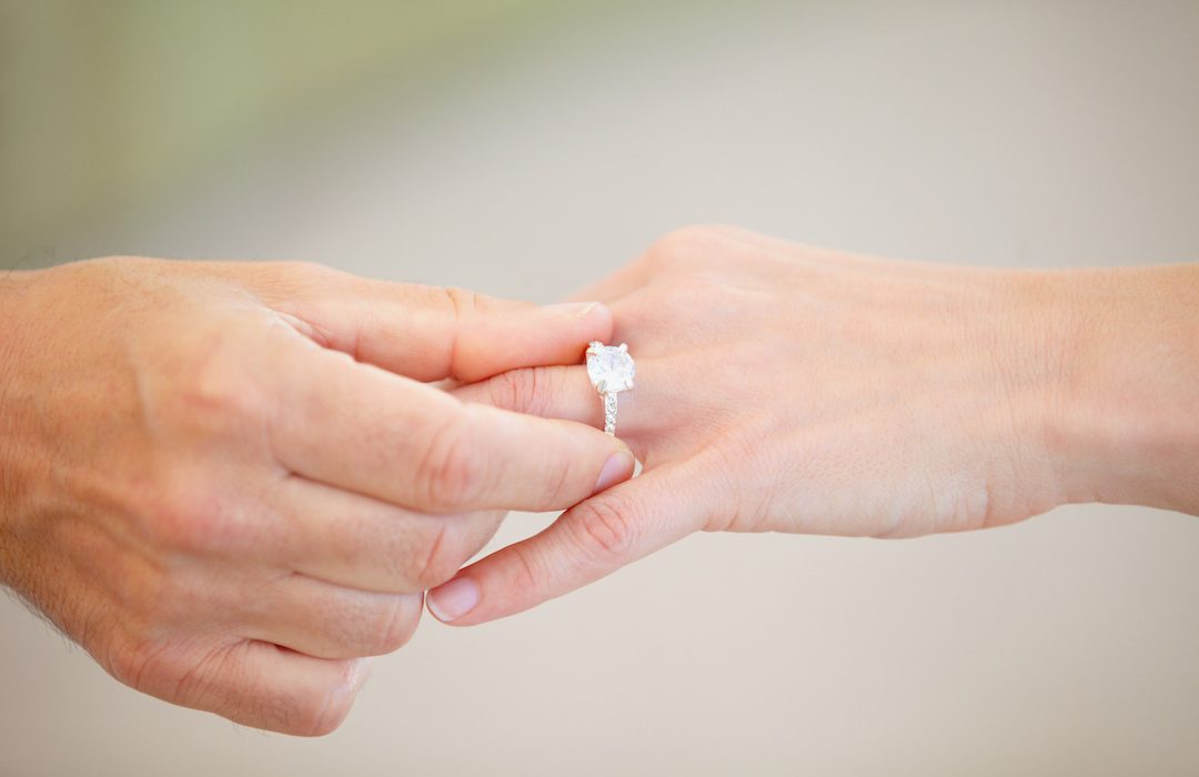 Cropped view of a mans hand sliding an engagement ring onto a womans ring finger.
