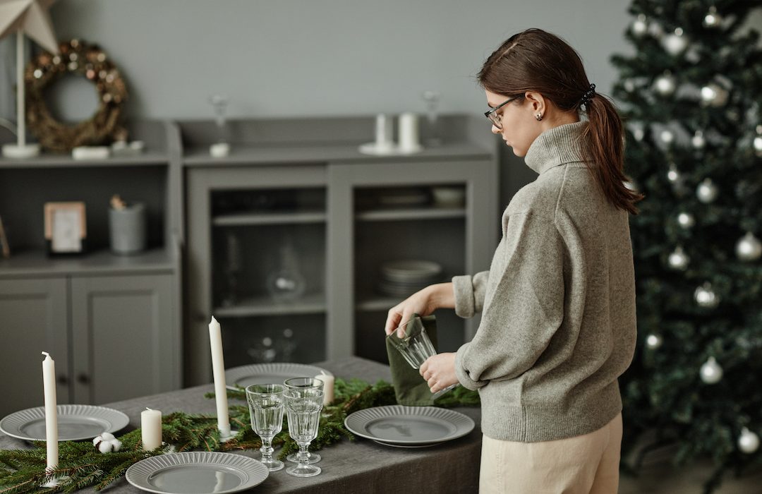 Side view portrait of young woman setting up plates on dining table decorated for Christmas with fir branches and candles in grey tones, copy space